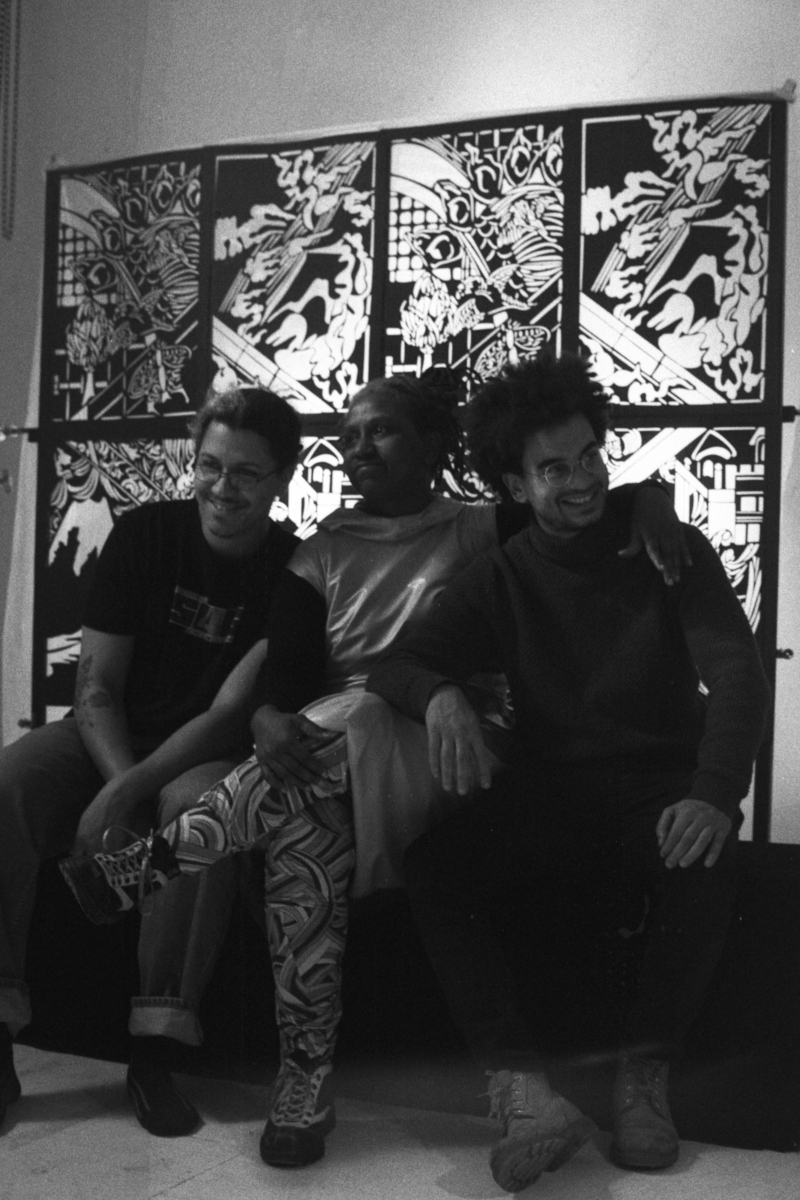 Three participants sit together smiling before the FableType light-cutting installation in a black-and-white behind-the-scenes photograph documenting the collaborative portrait session.
