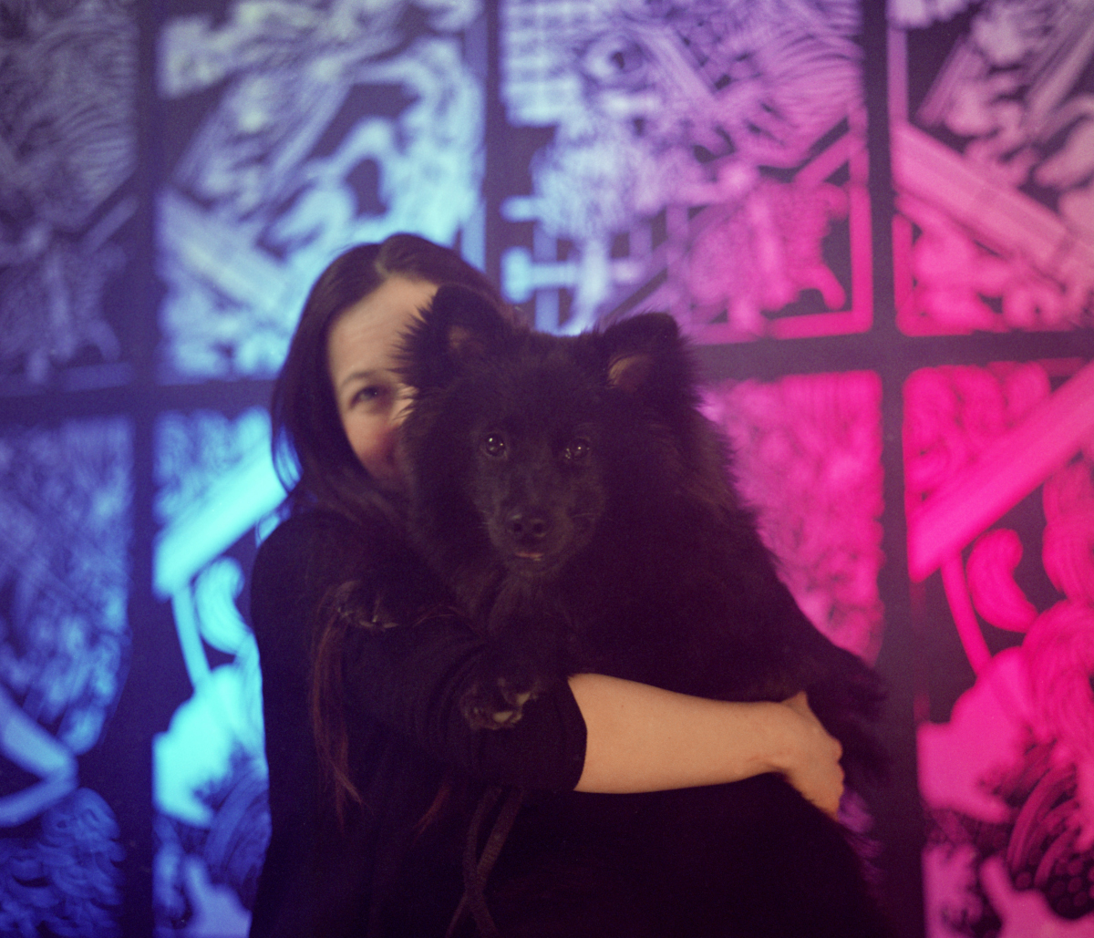 Portrait of a participant embracing a black dog before the illuminated FableType light-cutting installation, surrounded by blue and magenta light and soft atmospheric fog.
