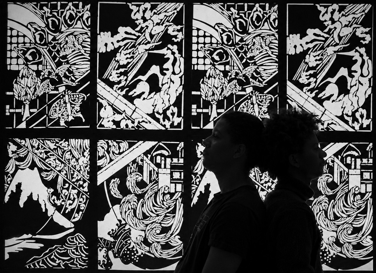 Two participants stand back-to-back in silhouette before the illuminated FableType light-cutting installation, emphasizing the graphic patterns and sculptural composition.
