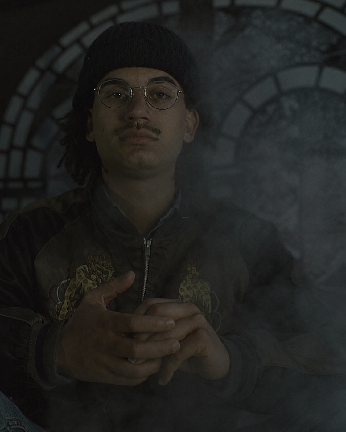 Portrait of a seated participant wearing glasses and a knit cap before an arched FableType light-cutting installation, with soft fog creating a quiet atmospheric setting.
