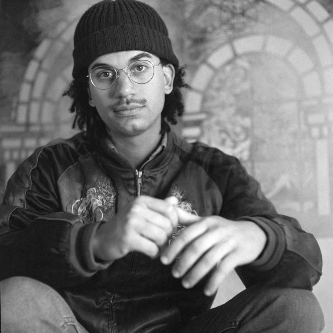 Black-and-white portrait of a seated participant wearing glasses and a knit cap before the FableType light-cutting installation, combining analogue portraiture with handcrafted light-cuttings.
