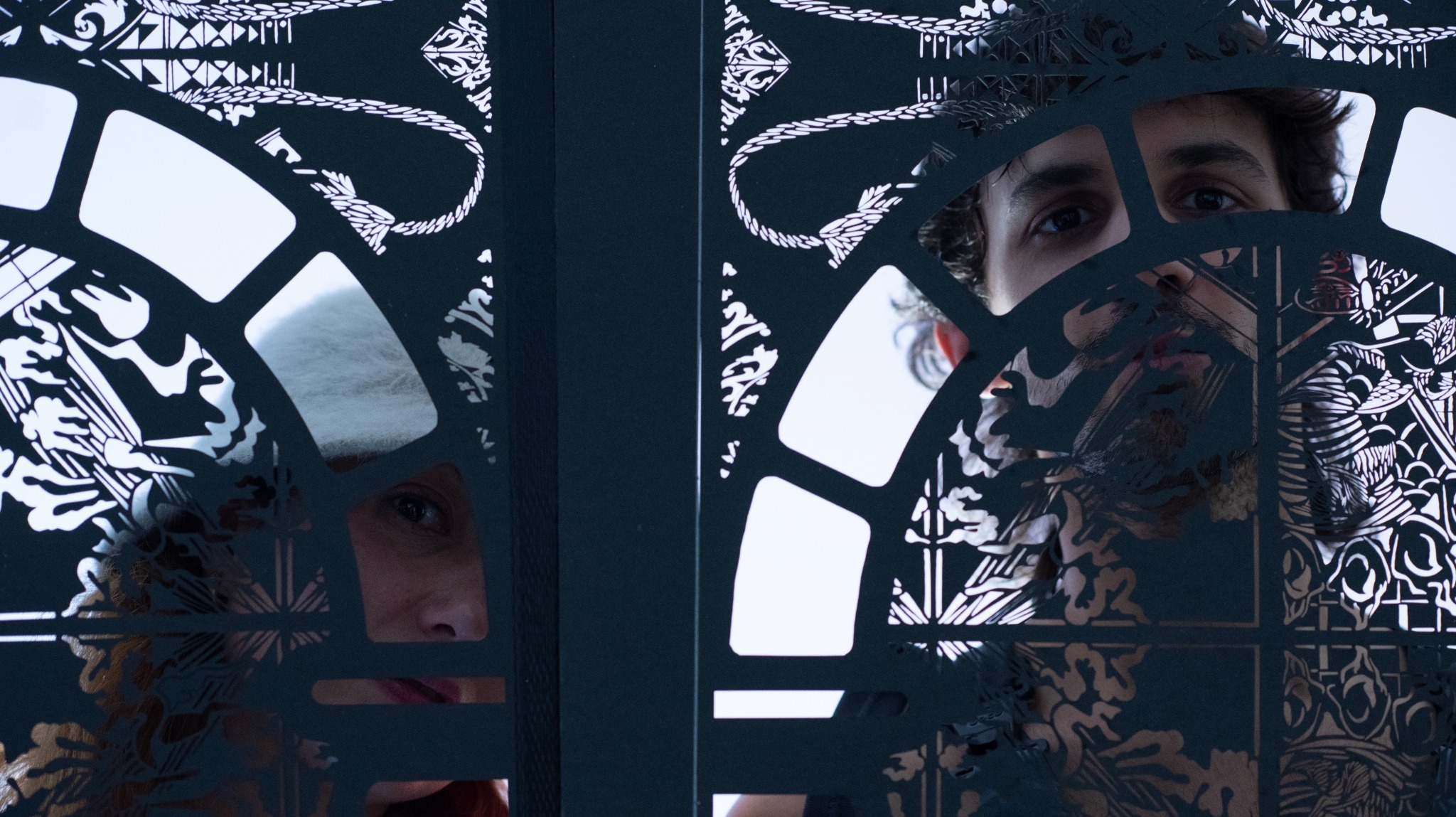 Two participants peer through the openings of a FableType light-cutting panel, their faces partially revealed by the intricate paper design.
