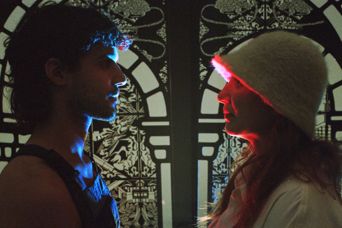 Two participants face one another in profile before the FableType light-cutting installation, illuminated by contrasting blue and red light in a narrative portrait.
