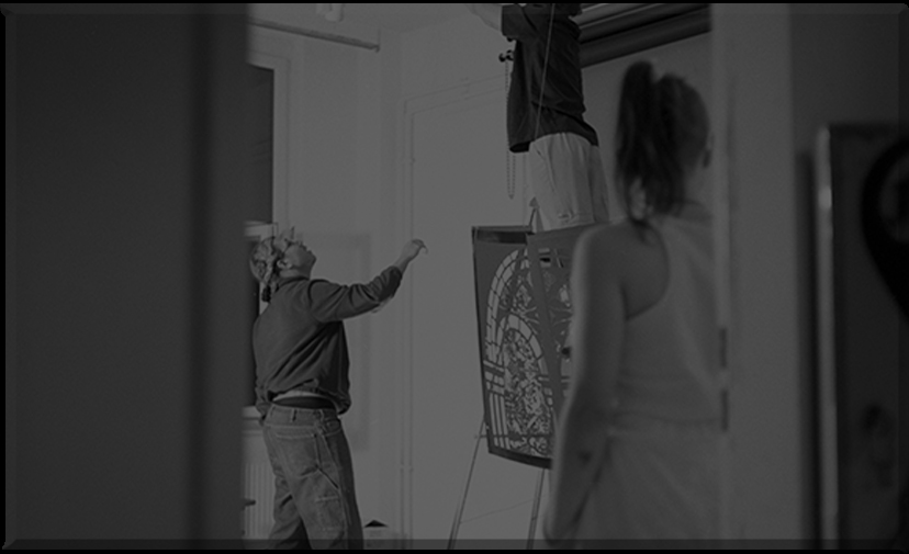 Behind-the-scenes photograph of the FableType installation being assembled, with participants positioning a light-cutting panel while another person works overhead preparing the exhibition.
