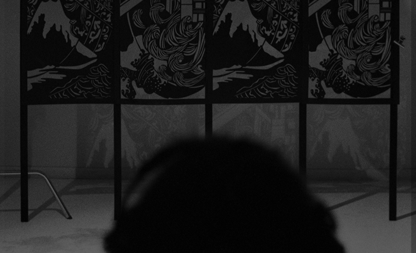 View of the FableType light-cutting installation from the audience, with the silhouette of a viewer in the foreground observing the illuminated paper artwork.
